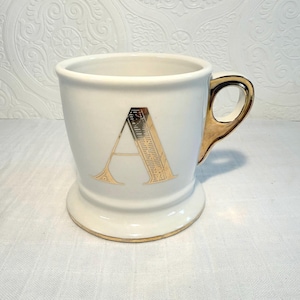 May include: White ceramic mug with a gold handle and a gold letter "A" on the front. The mug has a gold rim at the base and top. The mug is approximately 4 inches tall.
