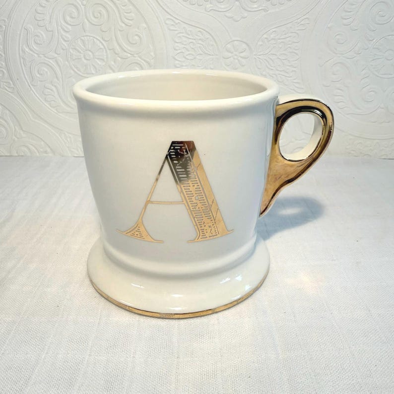 May include: White ceramic mug with a gold handle and a gold letter "A" on the front. The mug has a gold rim at the base and top. The mug is approximately 4 inches tall.