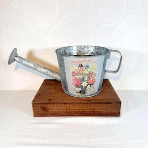 May include: A galvanized metal watering can with a floral design and the text "The Conard & Jones Co. West Grove, PA". The watering can has a handle and a spout. It is sitting on a wooden block.