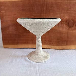 May include: A textured, off-white martini glass with a wide, triangular bowl and a slender stem. The glass has a rough, speckled surface and sits on a round base. The rim of the glass is dark green.