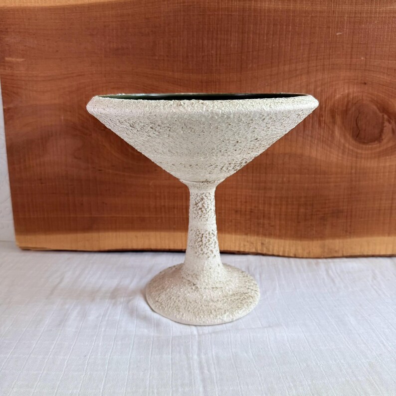 May include: A textured, off-white martini glass with a wide, triangular bowl and a slender stem. The glass has a rough, speckled surface and sits on a round base. The rim of the glass is dark green.