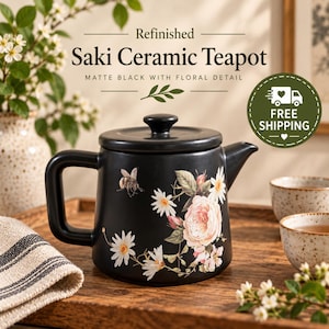 May include: A matte black ceramic teapot featuring a floral design with white daisies, a large pink rose, and a small bee. The text on the teapot reads Refinished Saki Ceramic Teapot, Matte Black with Floral Detail. It has a lid and handle.