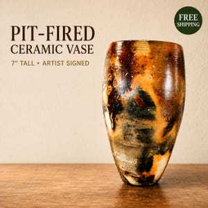 May include: A pit-fired ceramic vase with a mottled surface featuring shades of black, brown, and orange. The vase is 7 inches tall and is artist signed. The text on the image reads: PIT-FIRED CERAMIC VASE 7" TALL • ARTIST SIGNED.