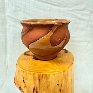 May include: A handmade ceramic bowl with a rounded shape and a wide opening. The bowl is a reddish-brown color with lighter brown, swirled accents. It sits on a natural wood base, showcasing the bowl's rustic charm.