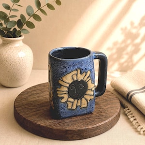 May include: A blue ceramic mug with a speckled finish and a square-shaped handle. The exterior features a raised, cream-colored sun motif with a dark, stylized face in the center. The mug has a sturdy, blocky design with a wide, flat base.