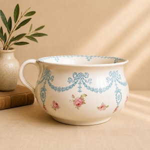 May include: Cream-colored ceramic chamber pot with a handle, adorned with light blue ribbon and floral patterns. Pink roses are scattered across the surface. A small vase with greenery is in the background.
