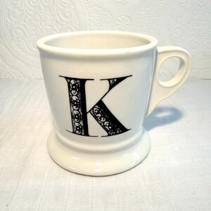May include: White ceramic mug with a black letter "K" in a decorative font. The mug has a handle and a pedestal base. The letter "K" is filled with a detailed, ornate design. The mug is likely for coffee or tea.