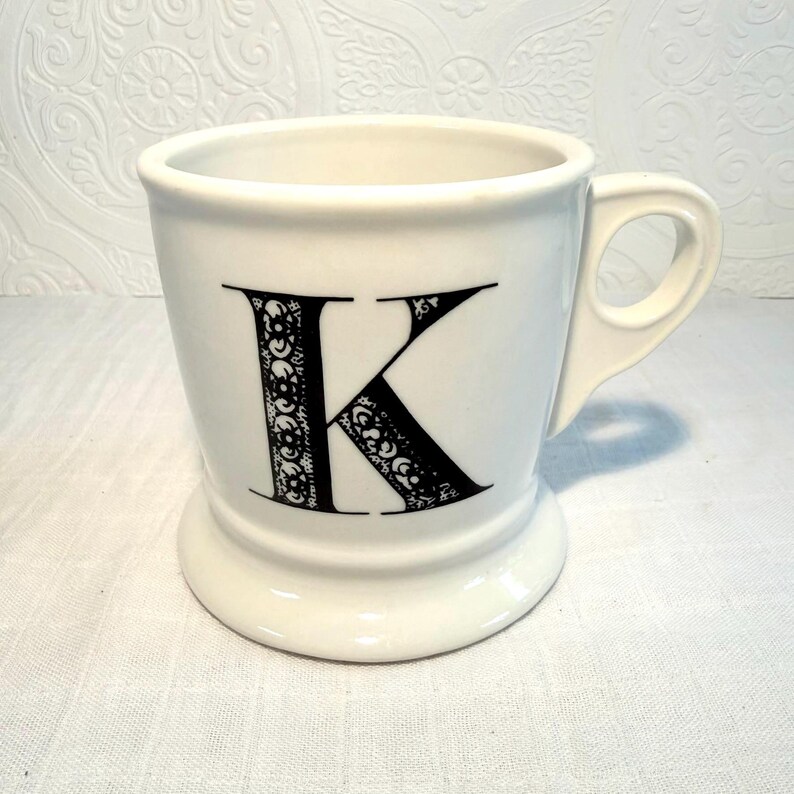 May include: White ceramic mug with a black letter "K" in a decorative font. The mug has a handle and a pedestal base. The letter "K" is filled with a detailed, ornate design. The mug is likely for coffee or tea.