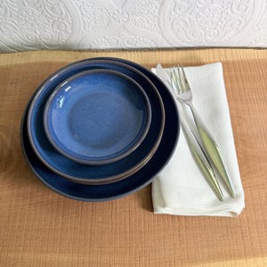 May include: A set of three blue ceramic plates with a dark rim, nested together. A white cloth napkin and two silver forks are placed to the right of the plates. The plates are on a wooden surface.