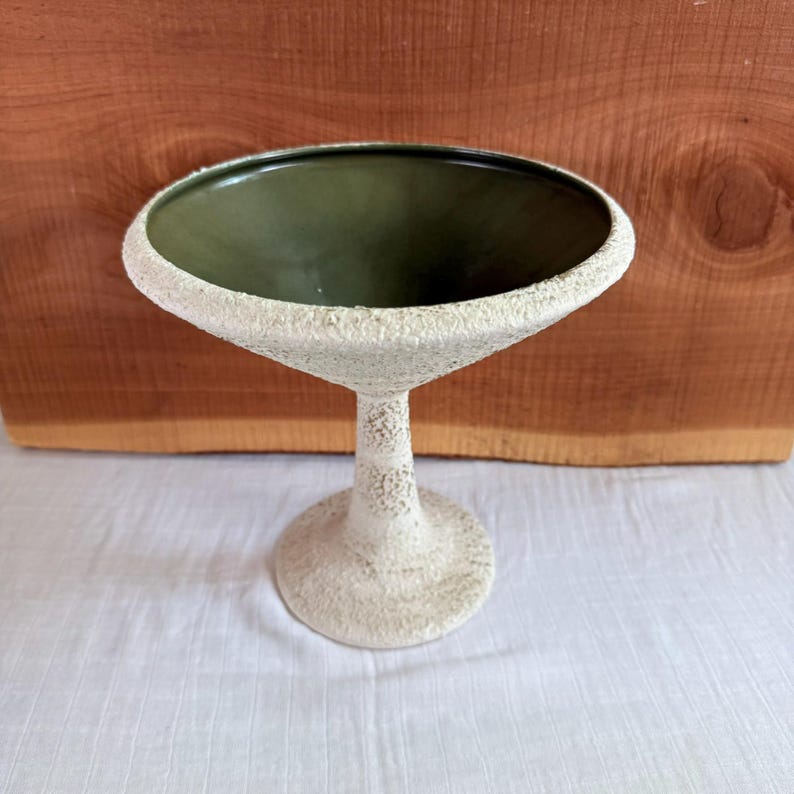May include: A textured, off-white ceramic pedestal bowl with a dark green interior. The bowl has a wide, flared rim and a textured stem that leads to a circular base. The background is a wooden surface.
