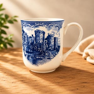 May include: White ceramic mug with a blue castle illustration. The mug features a curved handle and a detailed blue and white design of a castle scene. The mug is set on a wooden surface.