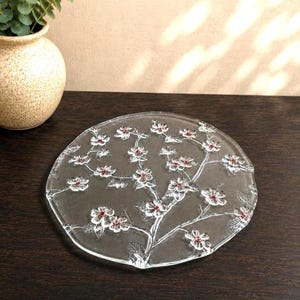 May include: A circular clear glass plate featuring an embossed branch design with multiple flowers. Each flower has a small, raised red center. The glass surface is smooth and transparent, showcasing the intricate floral pattern across the entire plate.
