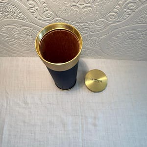 May include: A cylindrical container with a gold rim and a navy blue exterior. The interior is brown. A gold lid with the word "Guinness" is next to the container. The container is likely for a beverage.