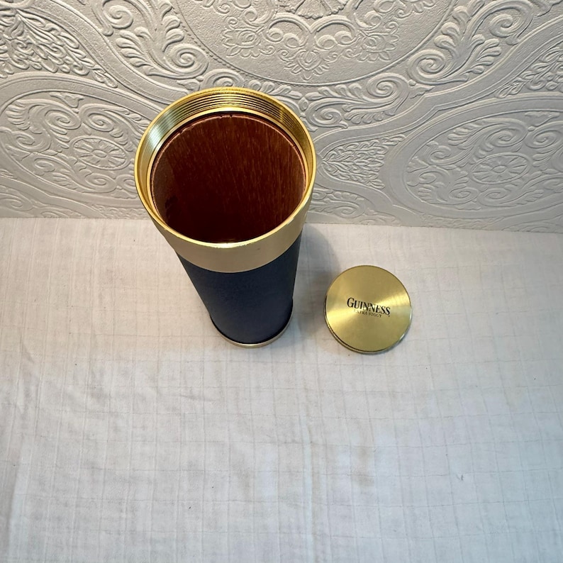 May include: A cylindrical container with a gold rim and a navy blue exterior. The interior is brown. A gold lid with the word "Guinness" is next to the container. The container is likely for a beverage.
