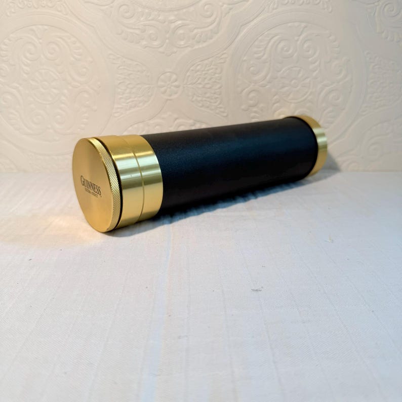 May include: A black and gold Guinness-branded travel bar kit. The cylindrical container has a black body with gold-colored metal accents on the ends and around the middle. The Guinness logo is visible on one end.