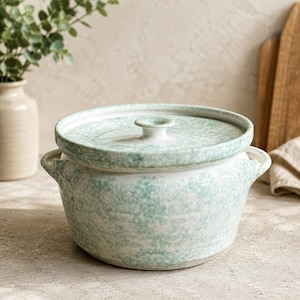 May include: A ceramic cooking pot with a lid. The pot is white with a speckled, light green pattern. It has two handles and a knob on the lid. The pot is designed for cooking and serving.