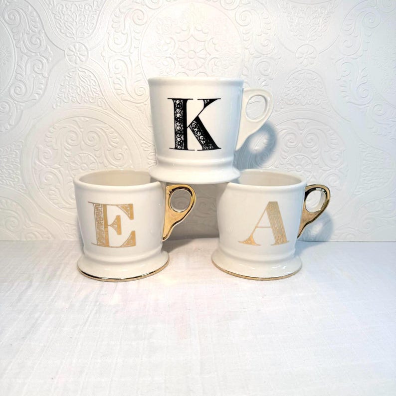 May include: Three white ceramic mugs with gold handles and gold lettering. The mugs are stacked, with the top mug displaying a black letter "K". The other mugs display the letters "E" and "A". The mugs have a decorative base and a gold trim.