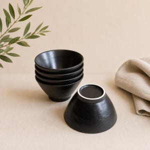 May include: A set of four black ceramic bowls with a smooth, matte finish. Three bowls are stacked vertically, while one bowl is placed upside down in the foreground, revealing a circular base with a white rim. The bowls have a conical shape.