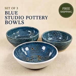 May include: A set of three blue studio pottery bowls with speckled glaze finishes. The bowls feature varying shades of blue, with one bowl displaying a white exterior base. The text on the image reads SET OF 3 BLUE STUDIO POTTERY BOWLS.