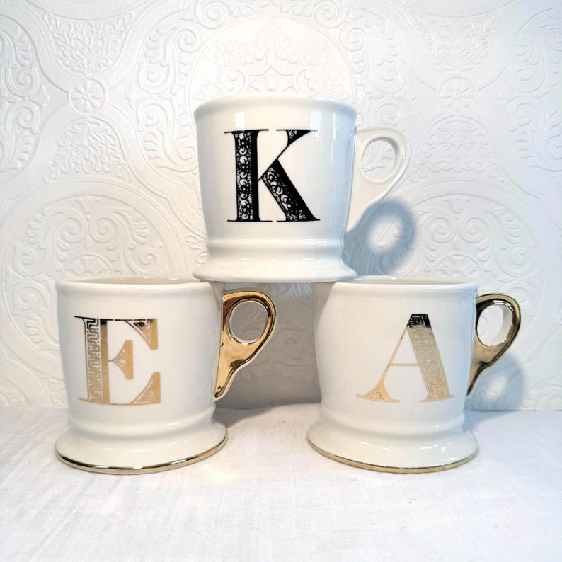 May include: Three white ceramic mugs with gold accents and large letter designs. The letters are black and gold, with the letters "K", "E", and "A" displayed on the front of each mug. The mugs have a classic shape with a rounded base and a handle.