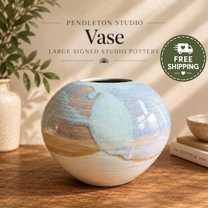 May include: A round ceramic vase featuring a multi-colored glaze with horizontal bands of white, tan, and light blue. The surface has a smooth, glossy finish with subtle texture. Text above the vase reads PENDLETON STUDIO Vase LARGE SIGNED STUDIO POTTERY.