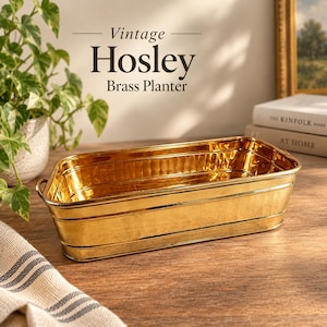 May include: A rectangular brass planter with a polished gold-tone finish, featuring horizontal ribbed detailing along the sides and small metal handles on each end. The planter is engraved with the text Vintage Hosley Brass Planter.