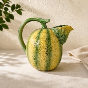 May include: A ceramic pitcher shaped like a cantaloupe melon. The pitcher is yellow with green vertical stripes and a green handle. A green leaf detail forms the spout. The pitcher sits on a light-colored surface.