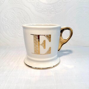 May include: A white ceramic mug with a gold handle and gold trim. The letter "E" is prominently displayed in gold on the front of the mug. The mug has a pedestal base.