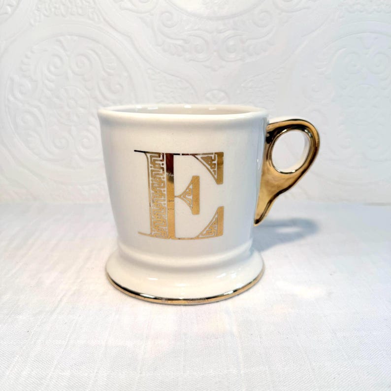 May include: A white ceramic mug with a gold handle and gold trim. The letter "E" is prominently displayed in gold on the front of the mug. The mug has a pedestal base.