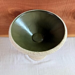 May include: A green ceramic pedestal bowl with a textured, off-white rim and base. The bowl's interior is a darker shade of green, and it sits on a short, rounded pedestal. The background is a wooden surface.