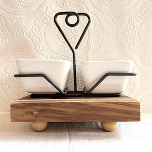 May include: A serving dish set featuring two white square bowls held by a black metal frame. The frame sits on a wooden base with small, round wooden feet. The background is a textured white wall.