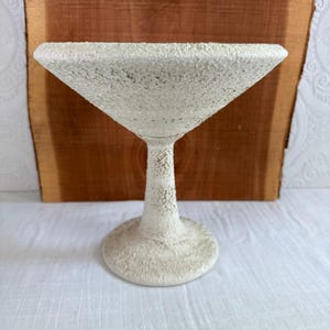 May include: A textured, off-white martini glass with a wide, triangular bowl and a slender stem. The glass has a rough, speckled surface, giving it a unique, handcrafted appearance. The base is round and sturdy. The background is a wooden surface.