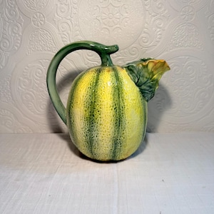 May include: A ceramic pitcher shaped like a cantaloupe, with green and yellow vertical stripes. The handle and spout are green, and a green leaf detail adorns the spout. The pitcher is on a white surface.