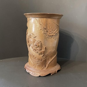May include: A light brown, cylindrical wooden vase with intricate carvings. The vase features a detailed relief of a lion and other figures amidst a landscape. The base has a decorative, scalloped edge.