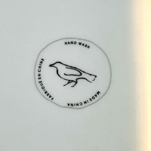 May include: Close-up of a white ceramic surface with a circular emblem. The emblem features a black bird illustration and the text "HAND WASH", "FABRIQU&Eacute; EN CHINE", and "MADE IN CHINA" in black lettering.