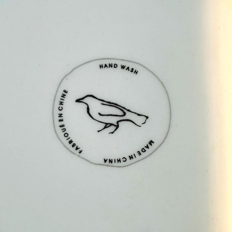 May include: Close-up of a white ceramic surface with a circular emblem. The emblem features a black bird illustration and the text "HAND WASH", "FABRIQU&Eacute; EN CHINE", and "MADE IN CHINA" in black lettering.