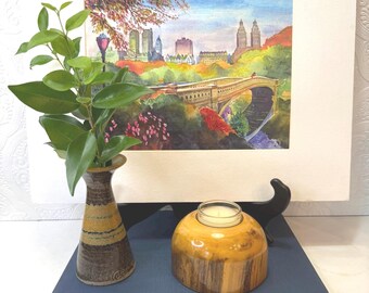 May include: A watercolor painting of a city park scene with a bridge and buildings, displayed on a white mat. A small ceramic vase with green leaves and a wooden candle holder with a lit candle sit on a blue book.