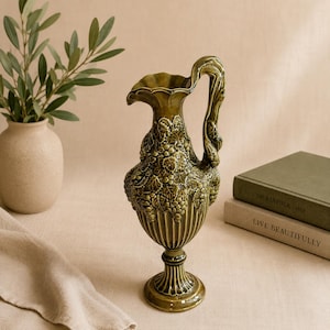 May include: A glossy olive green ceramic pitcher featuring a raised relief pattern of grape clusters and vine leaves on the body, a fluted base, and a curved handle. The pitcher has a scalloped rim and a tall, slender neck with a flared spout.