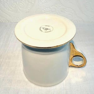 May include: A white ceramic mug with a gold handle and rim. The bottom of the mug has a white lid with a gold rim and a bird logo. The mug is on a white surface.
