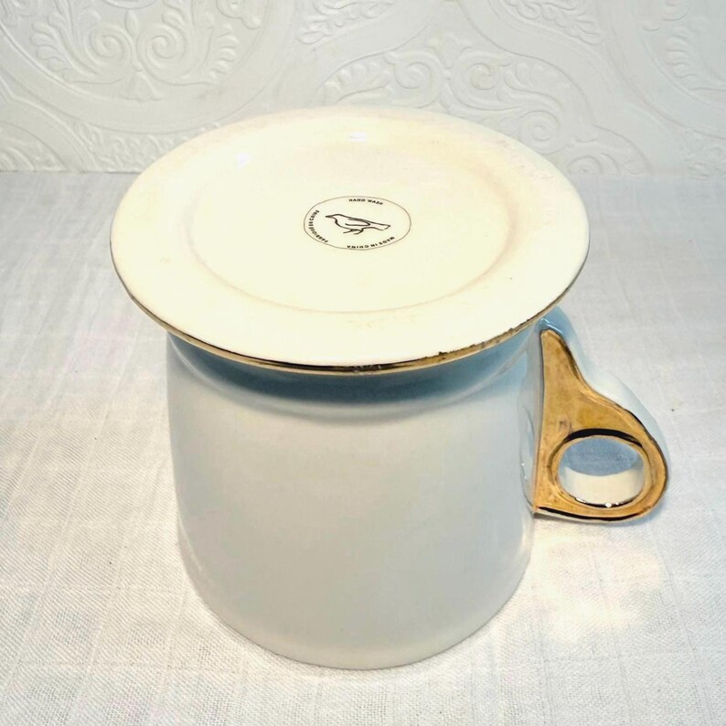 May include: A white ceramic mug with a gold handle and rim. The bottom of the mug has a white lid with a gold rim and a bird logo. The mug is on a white surface.