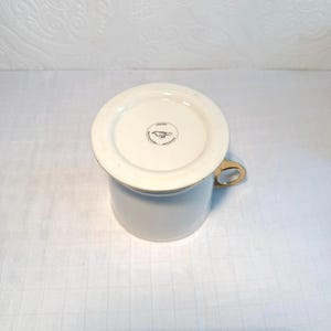 May include: A white ceramic mug with a gold handle and rim. The bottom of the mug is visible, featuring a circular emblem with a bird illustration. The mug is set against a white, textured background.