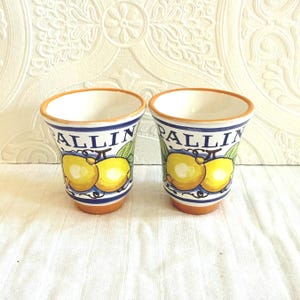 May include: Two ceramic cups with a white interior, featuring a design of yellow lemons and green leaves. The cups have a blue and white striped border and the word "PALLIN" is printed on the side. The rims and bases are a terracotta color.