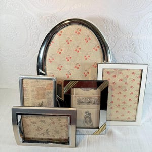 May include: A collection of decorative picture frames in various shapes and sizes. The frames have silver, white, and black finishes. The framed images feature vintage-style patterns with cherries and floral designs. The frames are arranged on a white surface.