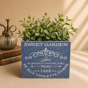 May include: A blue wooden planter box with the text "SWEET GARDEN" and other French text, filled with green leafy plants. A vintage-style brass plant mister and stacked books are in the background.