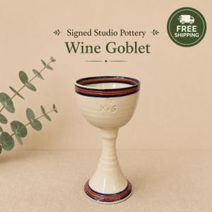 May include: A cream-colored ceramic wine goblet featuring a flared base and a stem with horizontal ridges. The rim and base are decorated with dark blue and reddish-brown bands. The letters KOG are embossed on the side of the bowl.