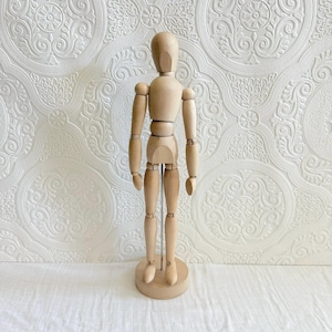 May include: A wooden artist's mannequin with articulated joints, standing on a round base. The mannequin is made of light-colored wood and is designed to be posed for drawing or sculpting. The background is a white wall with a textured pattern.
