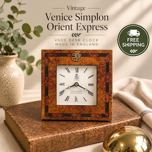 May include: A square desk clock with a burl wood frame and a white face featuring black Roman numerals. The clock face displays the text VSOE and Venice Simplon Orient Express. A gold-toned emblem is centered at the top of the frame.