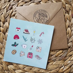 Peut inclure: Une carte de vœux bleu clair avec des illustrations sur le thème de Noël, dont un calendrier, des bottes, une flûte à champagne et un sucre d'orge. Une enveloppe marron avec une illustration estampillée est derrière la carte.