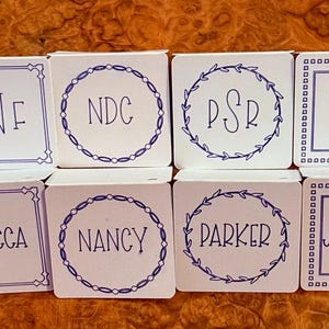May include: Six white square place cards with blue trim and lettering. The cards have different names and initials on them, including Rebecca, Nancy, Parker, Jacqueline, RNF, NDC, PSR, and JFM.