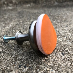 May include: A silver cabinet knob with a teardrop-shaped orange and white ceramic top.